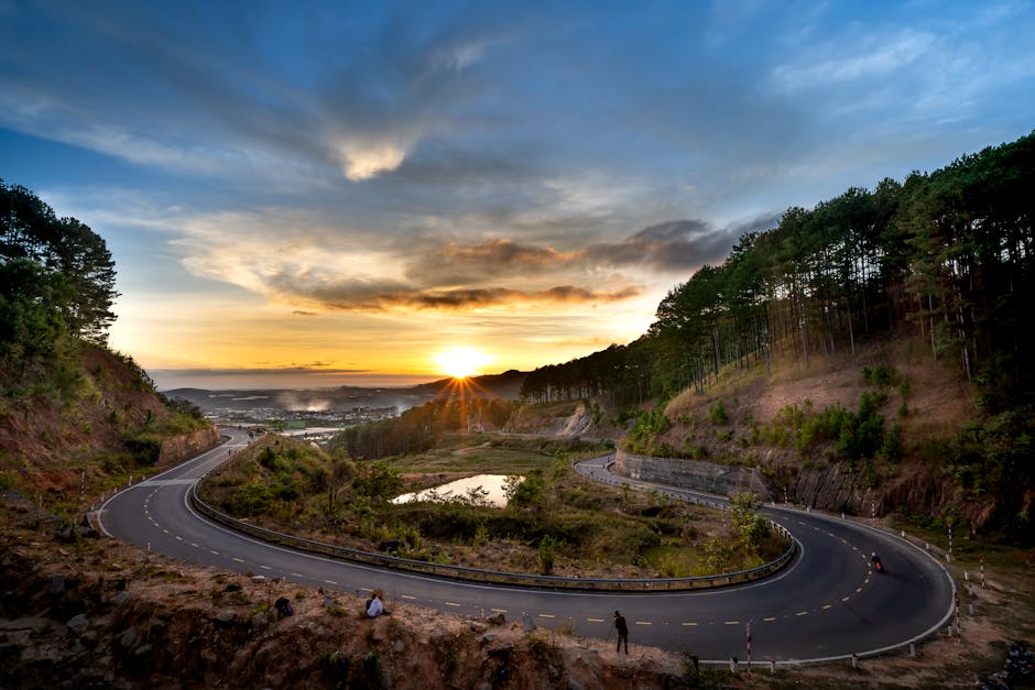 A winding road through the mountains at sunset, surrounded by lush forests and dramatic skies.